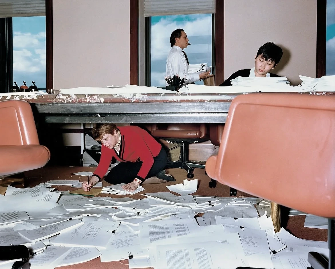 A conference room strewn with paper: one worker crawls beneath the table taking notes while colleagues sort stacks of documents above